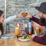 Pärchen am Tisch sitzend bildet mit abgerundeten Fingern ein Herz und hält ein knuspriges paniertes Fleischstück