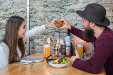 Pärchen am Tisch sitzend bildet mit abgerundeten Fingern ein Herz und hält ein knuspriges paniertes Fleischstück